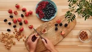 Overhead shot of fresh fruits, nuts, and hands preparing ingredients on a wooden board.